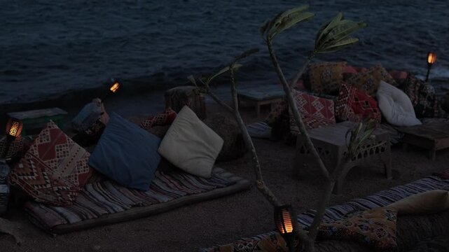 Bohemian beach lounge setup at dusk, cozy with soft glowing torch lamp, patterned cushions, rugs, low table, and small rocks on sandy shore beside dark ocean horizon