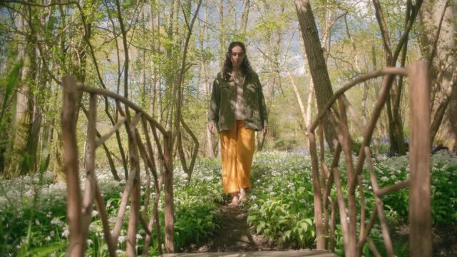 A woman walks through a woodland carpeted with white wild garlic flowers beneath a canopy of budding trees.