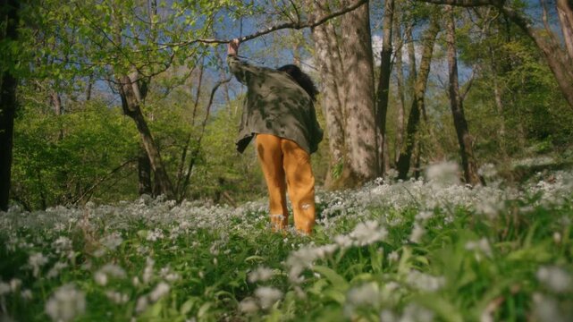 A wide shot of a woman walking through a field of wild garlic flowers beneath dappled woodland sunlight.