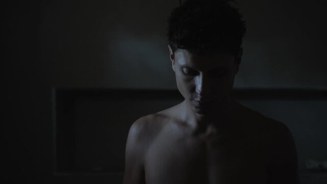 Young man lowers head from side profile to bowed pose in dim interior, cool rim light outlining face and upper body, introspective somber mood, near recessed niche on dark wall