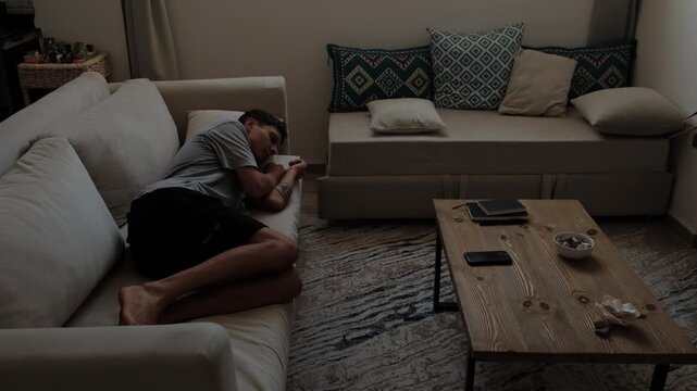 Man curled on light beige sofa resting with head on armrest beside low wooden coffee table with notebooks and smartphone in softly lit living room