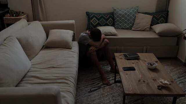 Young person sitting on floor between beige sofa and daybed, head bowed, arms wrapped around knees in dimly lit living room interior with subdued shadows and calm atmosphere
