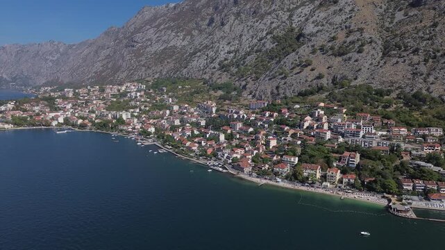 4K drone flight along coast of Bay of Kotor, Montenegro. Town with red tile roofs sits at foot of tall rocky mountains above deep blue water. Original unedited video, no color correction.