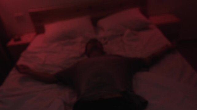 Depressed man sitting alone on his bed before falling back in emotional exhaustion and hopelessness, captured in slow motion with a Dutch angle and dark red low key cinematic lighting