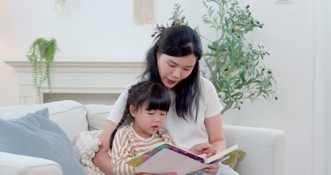 Asian single mother studies picture book with preschool daughter on sofa at home, supporting language learning, concentration, warm communication, literacy skill, bonding, child development daily