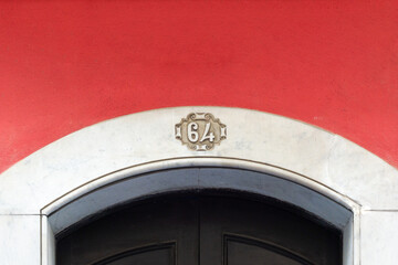 Ornate house number 64 appearing above an arched black doorway, set against a vibrant red plastered wall and a white marble archway in the city of Cádiz © photoopus
