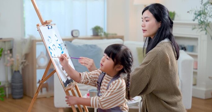 Asian single mother and preschool girl paint together near easel in soft daylight home, practicing brush skill, creative expression, close bonding, playful education, cognitive growth through art.