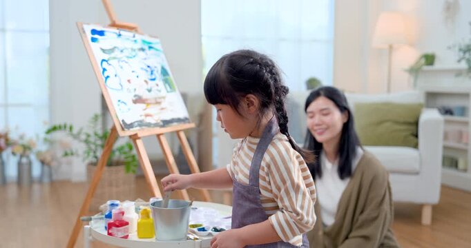 Little Asian girl stirs brush in paint tray beside easel while caring mother monitors progress in cozy home, supporting art practice, fine motor growth, concentration, creative learning, bonding.
