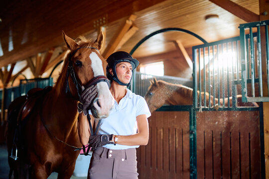 Equestrian rider wearing a helmet and gloves, standing with a saddled horse inside a bright stable looking ahead