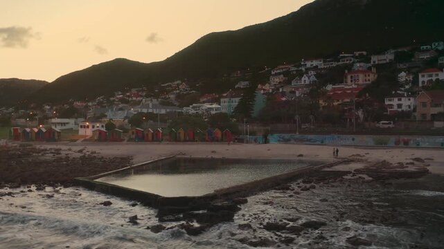 A scenic coastal landscape featuring a tidal pool along a rocky shoreline with hillside houses overlooking the calm ocean at beautiful golden sunset