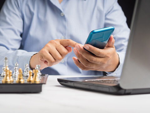 Businessperson using smartphone beside laptop and chessboard  