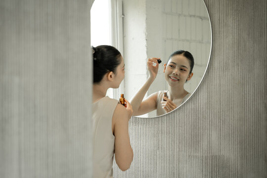 Young woman applies facial serum with a dropper while looking in a rou