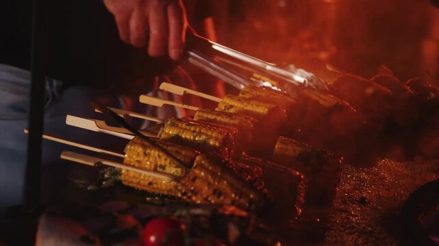 lose-up of corn on the cob on a stick sizzling on a hot griddle beside an open fire while a person turns it with metal tongs. Outdoor cooking with flames and a rustic barbecue atmosphere.