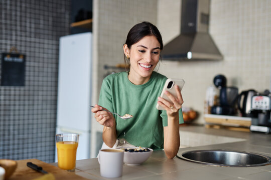 Portrait of a young woman preparing and eating breakfast, drinking juice and using a mobile phone smartphone  in the kitchen at home