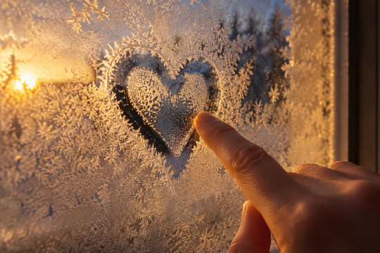 Finger tracing a heart shape through frost on a window at golden winter sunset