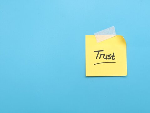 Yellow adhesive note with handwritten text trust taped on blue background with copy space. Conceptual image for business reliability, confidence, honesty, commitment, and communication message
