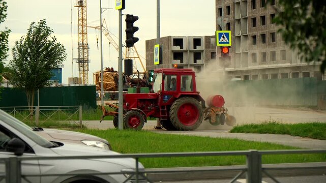 Red tractor cleans city sidewalk beside active construction zone creating thick clouds of dust. Utility vehicle sweeps paved road during urban maintenance work under daylight conditions.