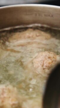 Savory meatballs simmer in bubbling hot broth inside stainless steel cooking pot. Metal ladle stirs boiling soup slowly, preparing nourishing homemade meal with fresh culinary ingredients.
