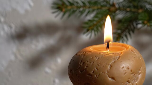 Textured ochre sphere candle glows beneath an evergreen sprig against a pale patterned wall, revealing a recessed wax pool and rugged surface in a quiet Christmas close-up with space on the left side