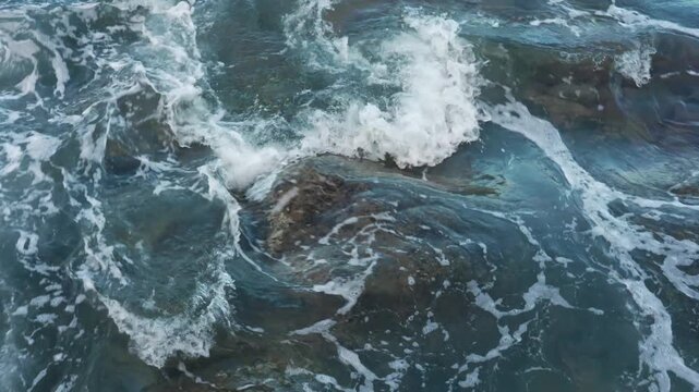 Gentle waves ripple across a clear blue ocean, revealing the faint outline of a submerged rock beneath the surface as sunlight dances through the water. The soft motion creates shifting patterns.