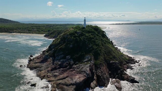 Farol das Conchas rises above a rocky headland, beaches and Atlantic waters on Ilha do Mel, Paranagua, Parana, Brazil, during a drone forward flyover.
