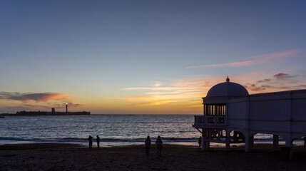 People enjoying a tranquil sunset on La Caleta beach in Cádiz, observing the historic Balneario de La Palma y del Real building and the distant San Sebastián Castle lighthouse © photoopus