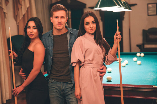 One young man stand beyween two models. They look on camera seriously. Young women hold billiard cues in hands. They stand in playing room.