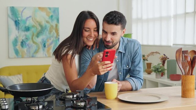 A cheerful duo gathers at a bright kitchen counter, peering at a red smartphone while smiling and sharing a light moment. Nearby, a mug, plate, and cookware accent the cozy home scene.