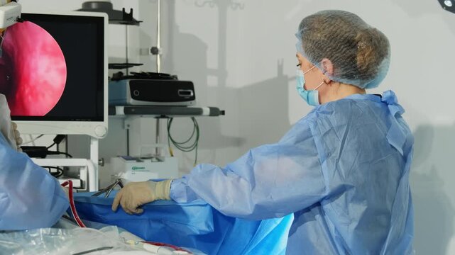 Female surgeon in gown and mask controls patient condition as colleague work. Woman doctor takes part in complicated surgery at hospital