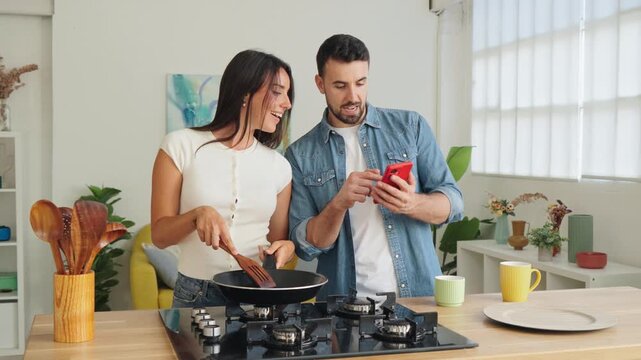 Two people share a light hearted moment in a clean kitchen as one stirs a pan while the other glances at a phone. Natural daylight, polished surfaces, and a tidy counter create a friendly home