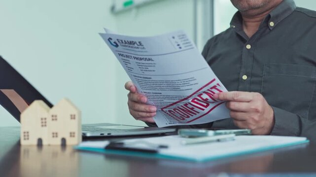 Businessman holding confidential project proposal paper beside laptop computer small wooden house model at office desk.