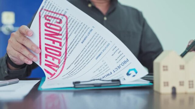 Businessman sitting indoors reviews confidential real estate agreement papers beside wooden house models while handling legal property documentation.