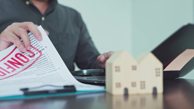 Businessman reviewing stamped foreclosure document on desk beside wooden house models laptop in office.