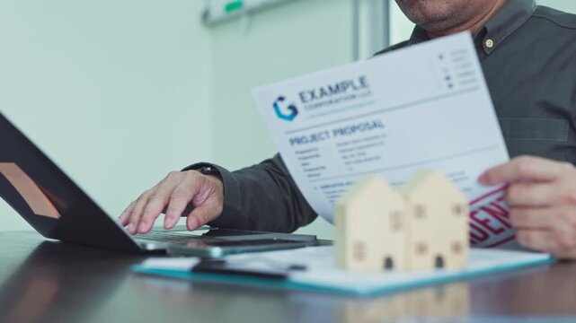 Real estate investor reviewing project proposal document beside house models typing laptop keyboard in office.