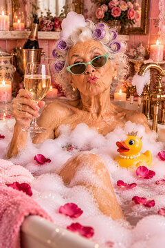 Stylish senior woman enjoying a luxurious bubble bath with champagne, hair curlers, sunglasses, rose petals, and a rubber duck in a rich vintage bathroom.
