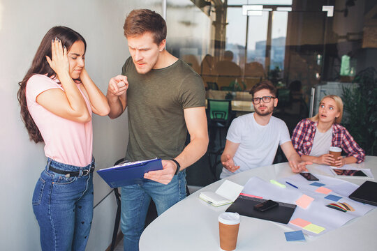 Angry young amn hold plastic tablet woth papers and point on it. He is angry and screaming. Two other people sit behind them. They are amazed and scared. Brunette keep ears and eyes closed.