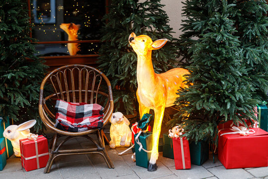 Magical winter display featuring an illuminated deer figure, glowing rabbits, red gift boxes, and a cozy wicker chair beside pine trees. Christmas outdoor festive decor