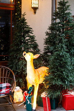 Festive christmas display with illuminated deer figure, glowing rabbits, red gift boxes, and a cozy chair beside pine trees with lights. Magical winter Christmas street decoration concept.
