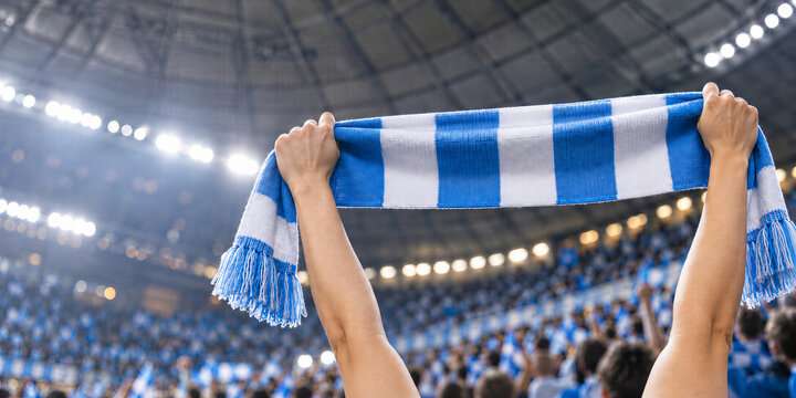 Sports fan holds blue and white striped scarf overhead in a crowded stadium. Football match support and team spirit. Live sports event and spectator culture. Fan enthusiasm and celebration