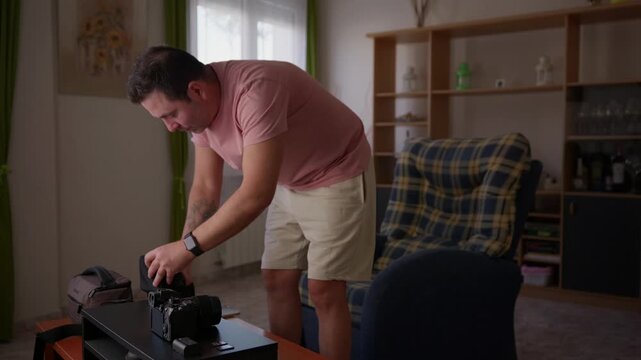 A man bends over a table to handle and prepare camera equipment in a living room showing a practical moment of audiovisual production.