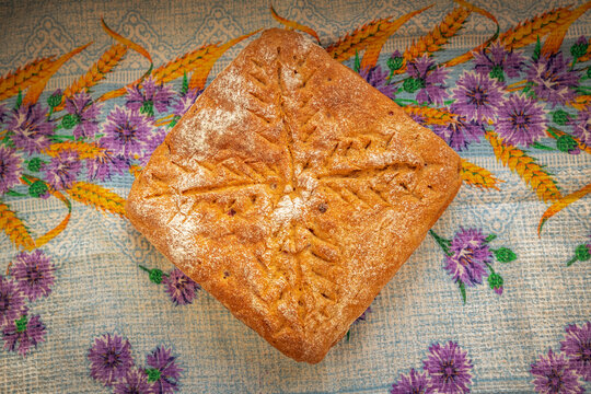 Golden-brown homemade loaf cooling on a patterned kitchen cloth, with a flour-dusted crust and decorative cuts arranged in a branching wheat-like design