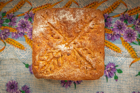 Rustic homemade bread loaf with a golden-brown flour-dusted crust and decorative wheat-like scoring, resting on a colorful patterned kitchen cloth