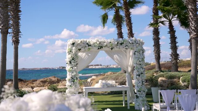 A wedding setup takes place near the sea with white flowers framing an arch. Chairs are arranged in front of the arch. Palm trees sway under a bright sky.