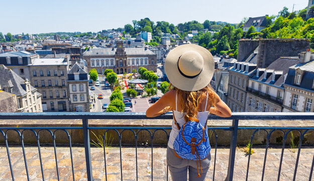 Young female traveler with straw hat and backpack looking at panoramic view of historic city Morlaix in Brittany France
