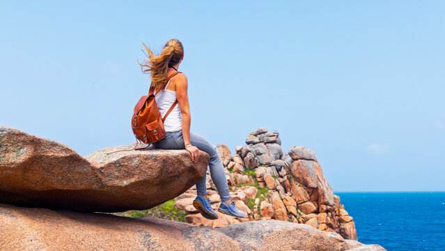 Young female hiker with leather backpack sitting on pink granite rock cliff overlooking ocean coast in Perros-Guirec Brittany France