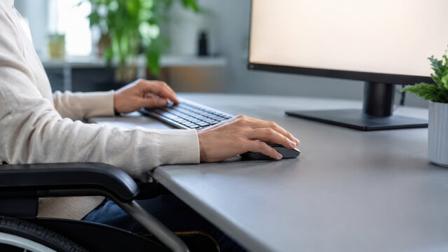 Wheelchair user engaged in computer activity at modern desk, working with keyboard and mouse in bright office environment