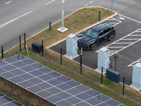 Aerial view of an electric vehicle charging station with a black car plugged in, adjacent to a solar panel array in the United Kingdom.
