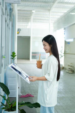 Asian teen girl reads the menu while standing at the entrance to the establishment while holding iced coffee drink