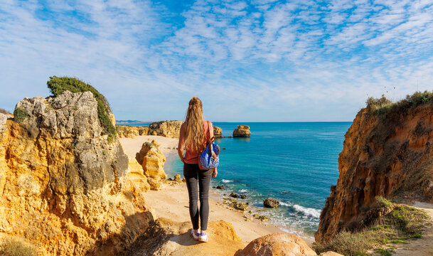 Young female traveler with backpack standing on cliff looking at beach and ocean in San Rafael Algarve Portugal