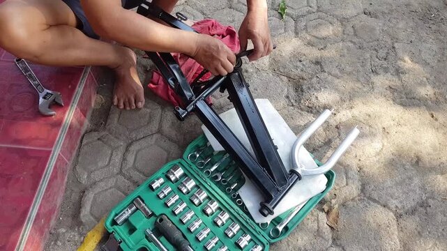 A man crouching outdoors assembling used bicycle frame with toolkit collection and socket set on paving block for restoration project